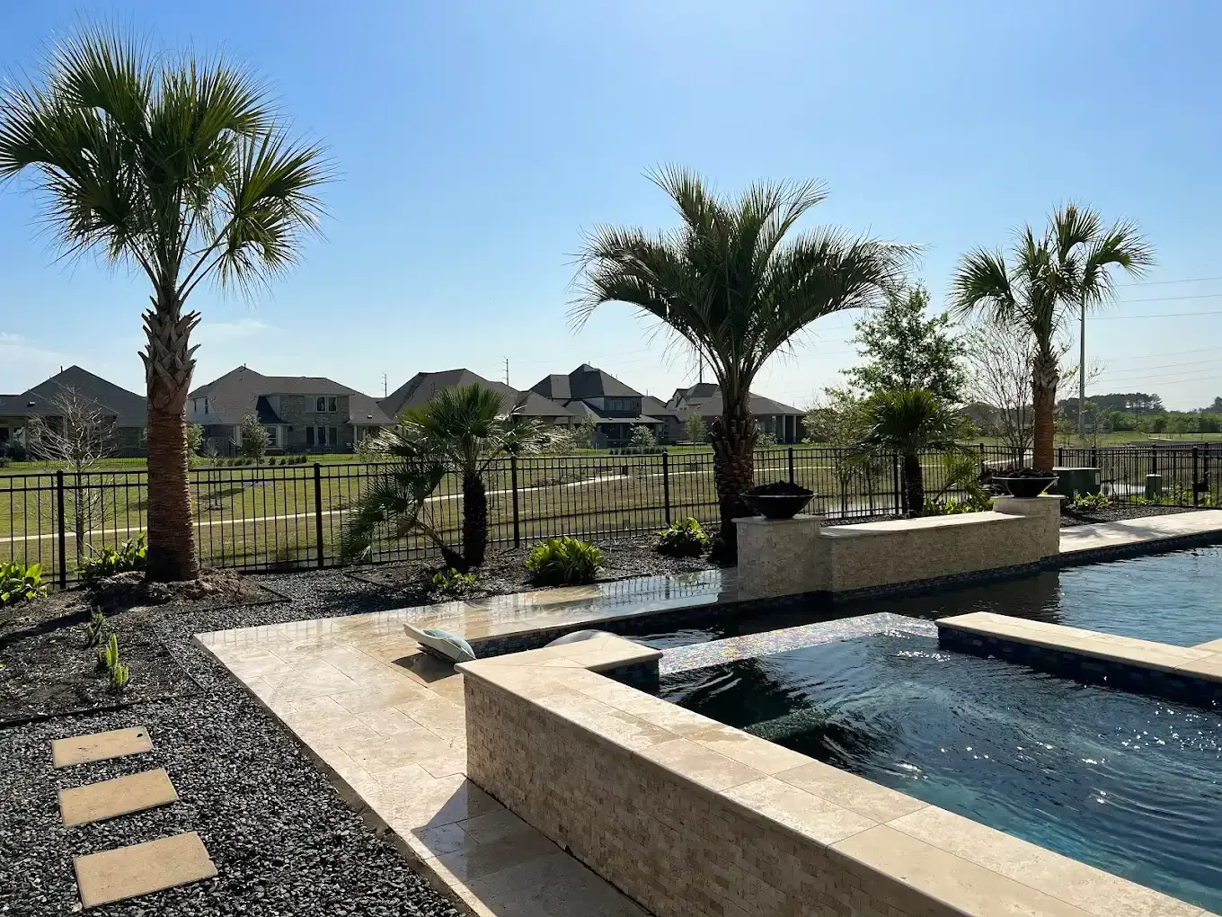 Outdoor pool area with palm trees, stone patio, and distant houses on a sunny day.
