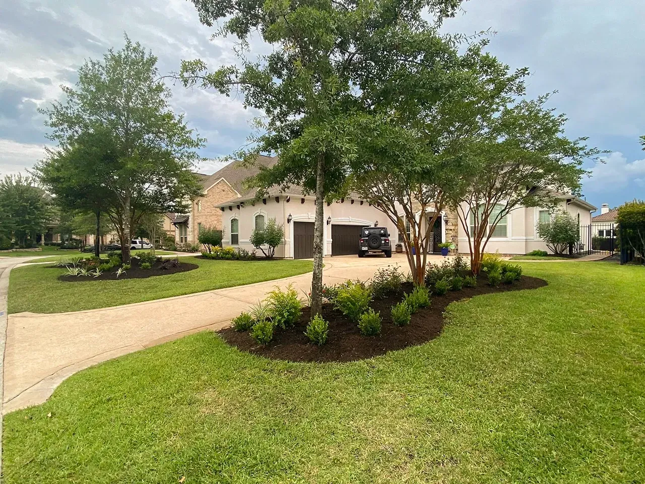 Suburban house with driveway, green lawn, and landscaped trees under a cloudy sky