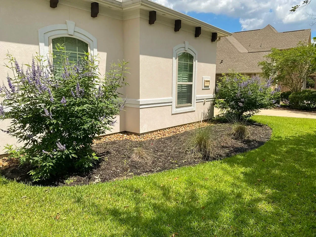 Single-story cream house with arched windows, green lawn, and purple flowering shrubs.