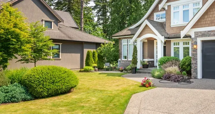 Two-story houses with green lawns, trees, and shrubs. One has a stone facade and driveway.
