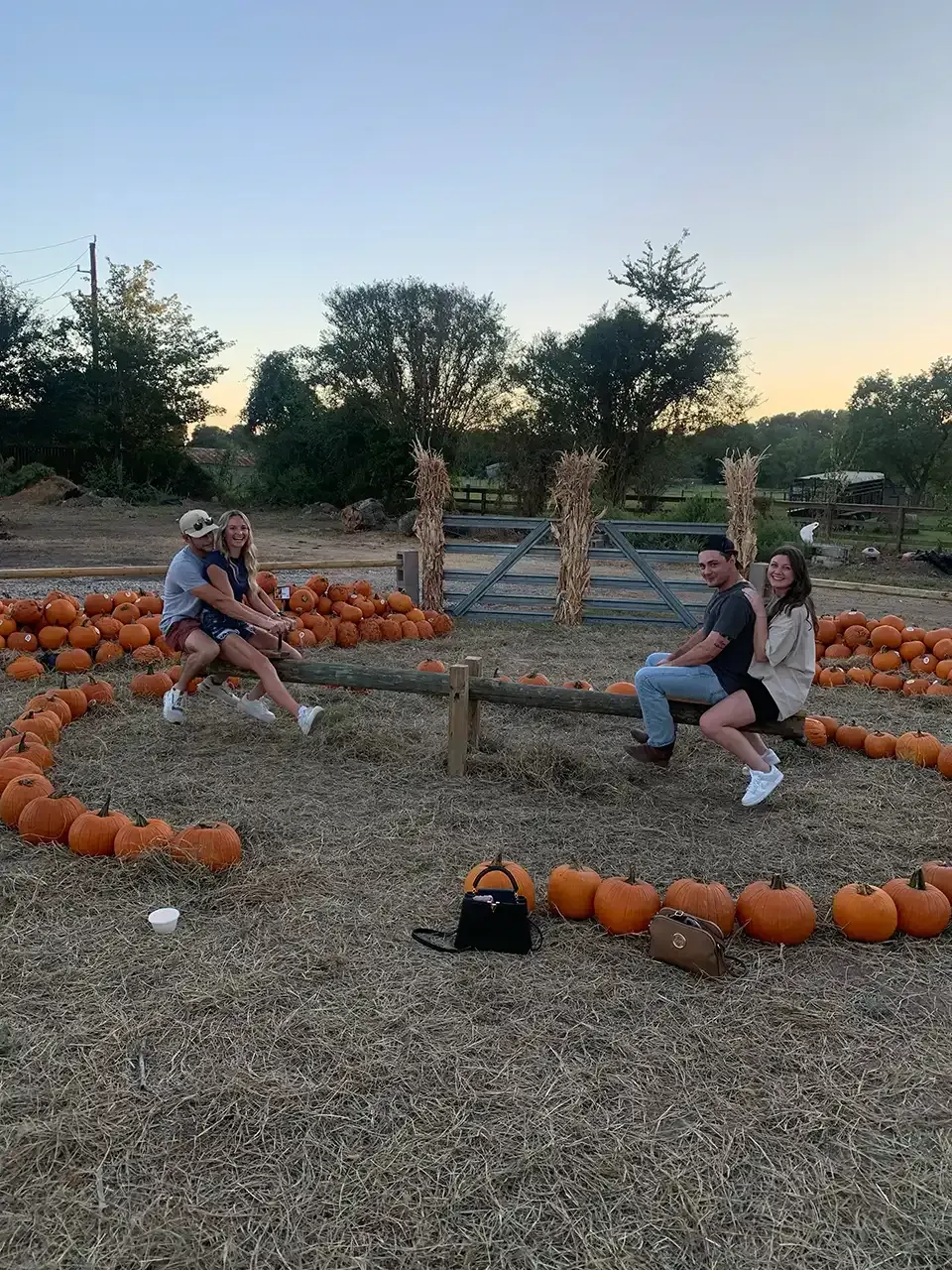 Two couples on a wooden seesaw in a pumpkin patch, surrounded by pumpkins in a heart shape at dusk.