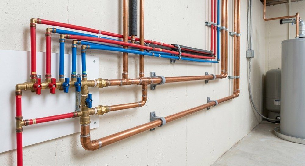 Color-coded plumbing pipes and valves mounted on a white basement wall, beside a water heater.