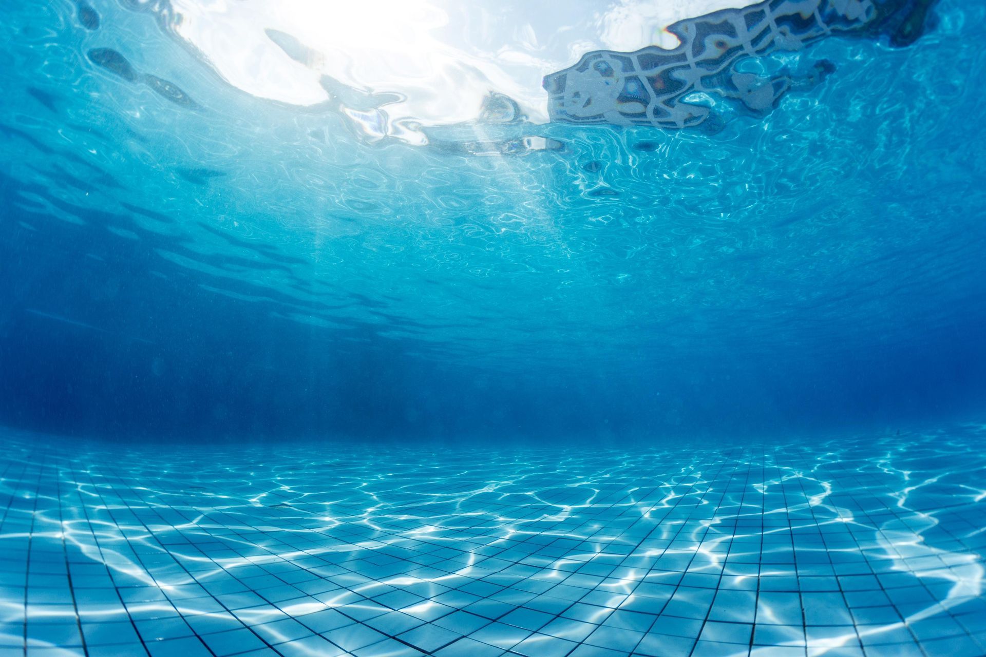 Underwater view of a swimming pool. Sunlight ripples on the sandy bottom and blue water.