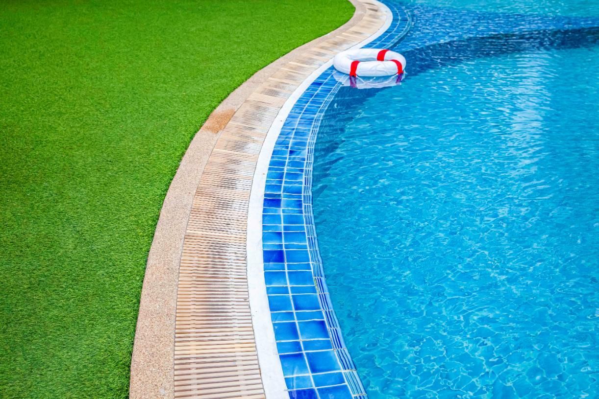 Poolside with turquoise water, blue tile edge, tan border, and green turf. A life preserver floats in the pool.