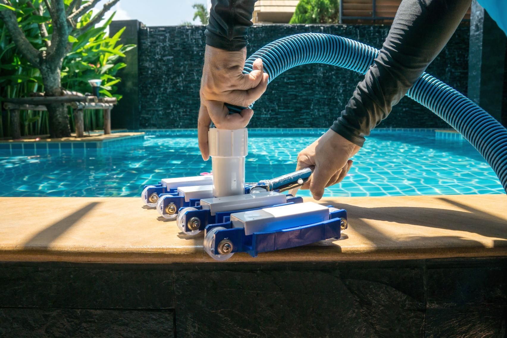 Person attaching a pool vacuum hose to a blue and white vacuum head next to a pool.