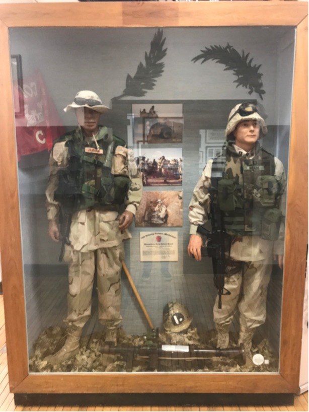 Display case with two mannequins in military uniforms, photos, and a helmet. Brown frame.