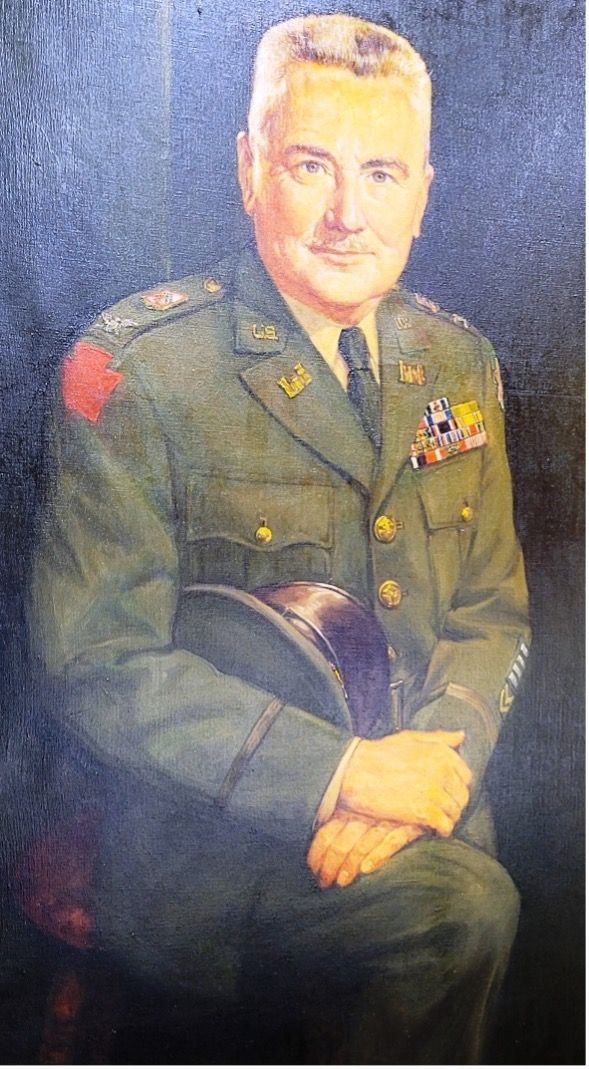 Portrait of a man in military uniform, holding hat. Red shoulder patch. Dark background.
