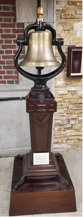 A bronze bell on a wooden stand with a brick and stone background.