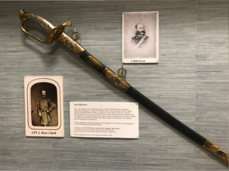 Display of a saber, portraits, and text panels. Saber has a gold hilt and black grip, flanking portraits and descriptive text.