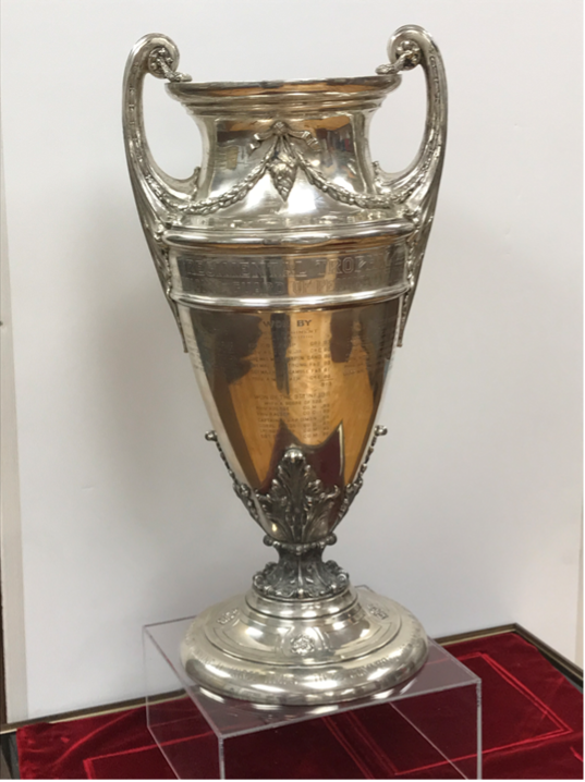 Silver trophy cup with handles, displayed on a clear stand over a red surface.