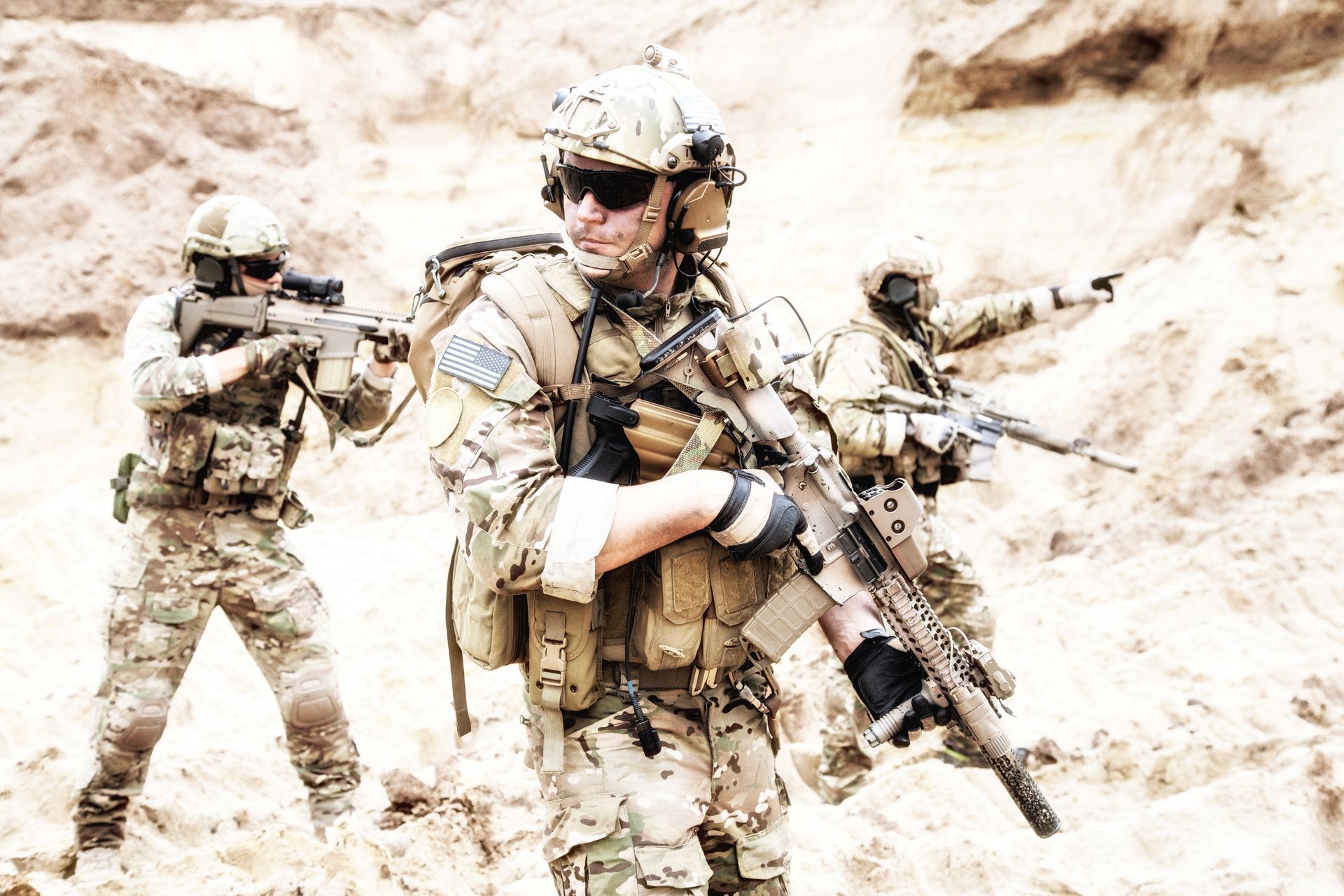 Three soldiers in tan camouflage gear with weapons, outside.