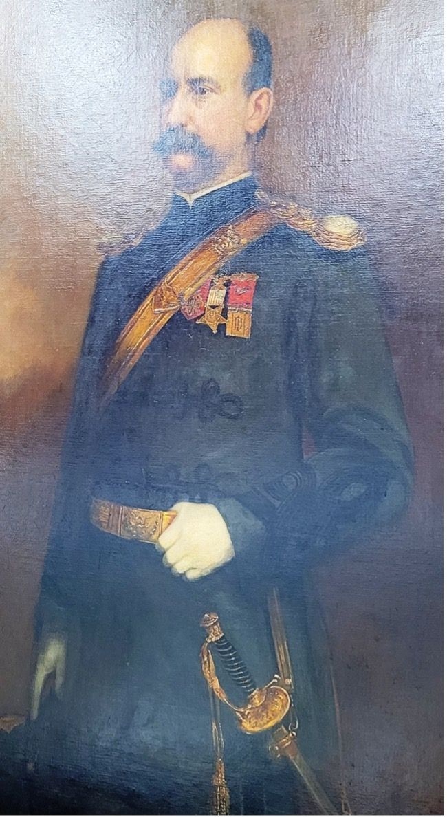 Man in dark military uniform with gold sash, medals, and sword, looking ahead.
