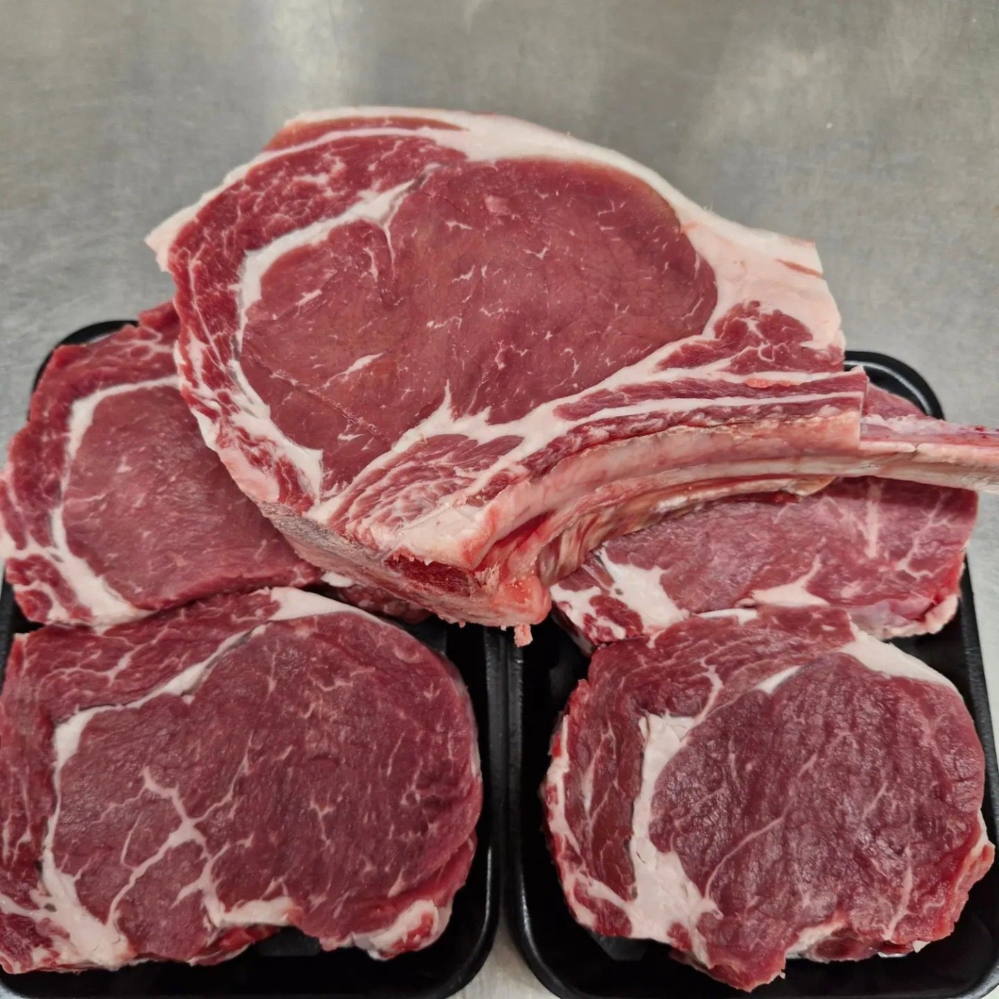 Raw ribeye steaks arranged in black trays, showing marbling and bone.