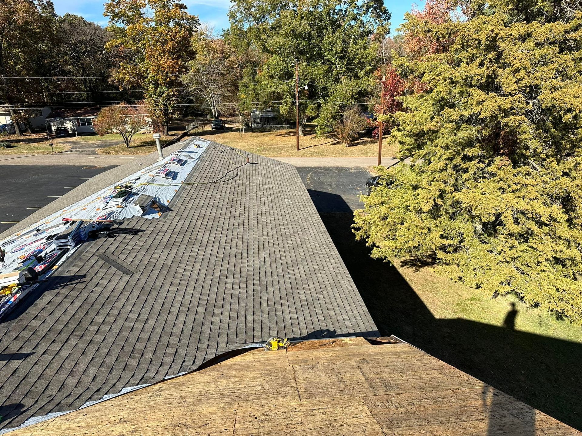 Roof partially stripped, asphalt shingles visible. Trees and a parking lot are in the background on a sunny day.