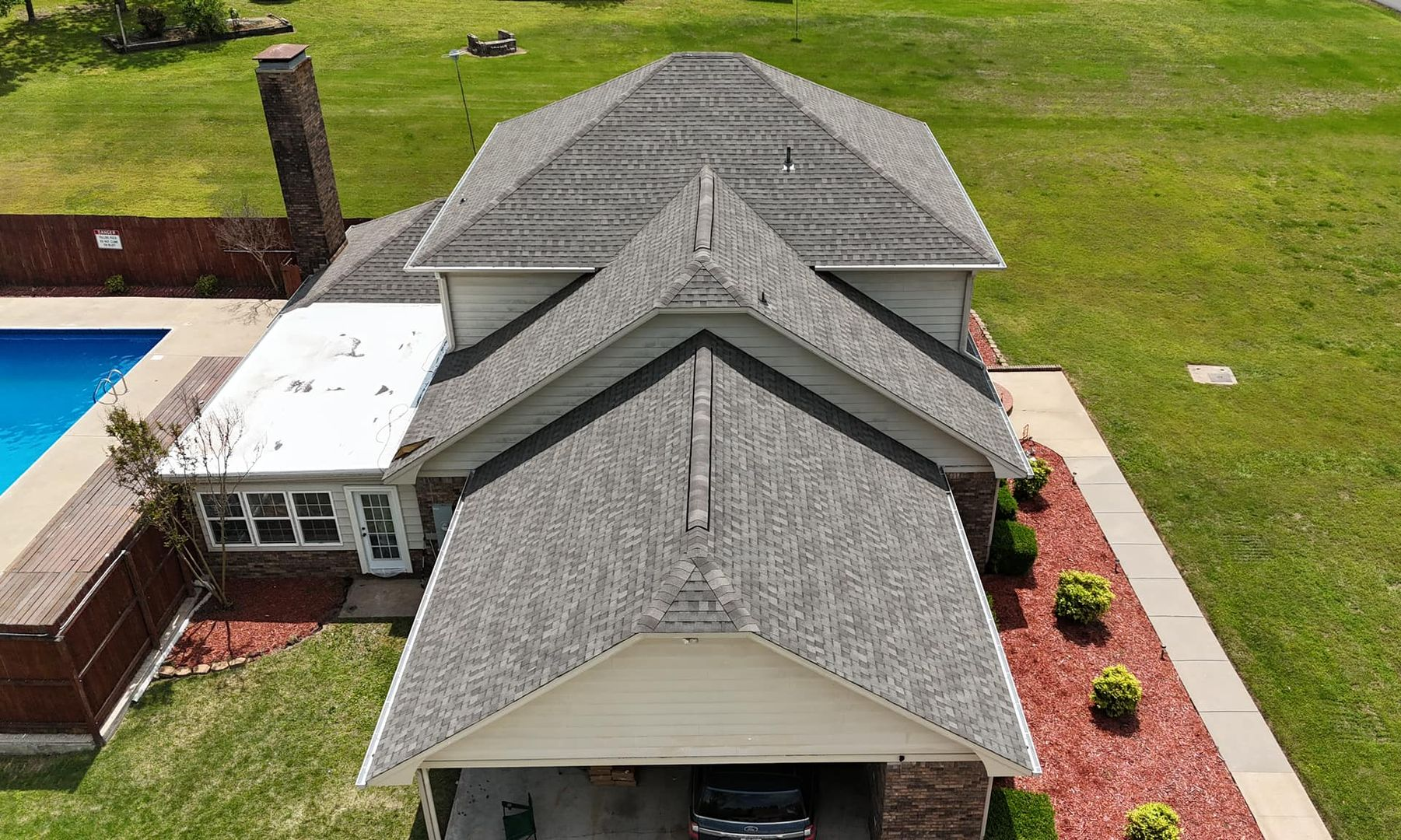 Overhead view of a two-story house with a gray shingle roof, chimney, and a carport. A pool is to the left.