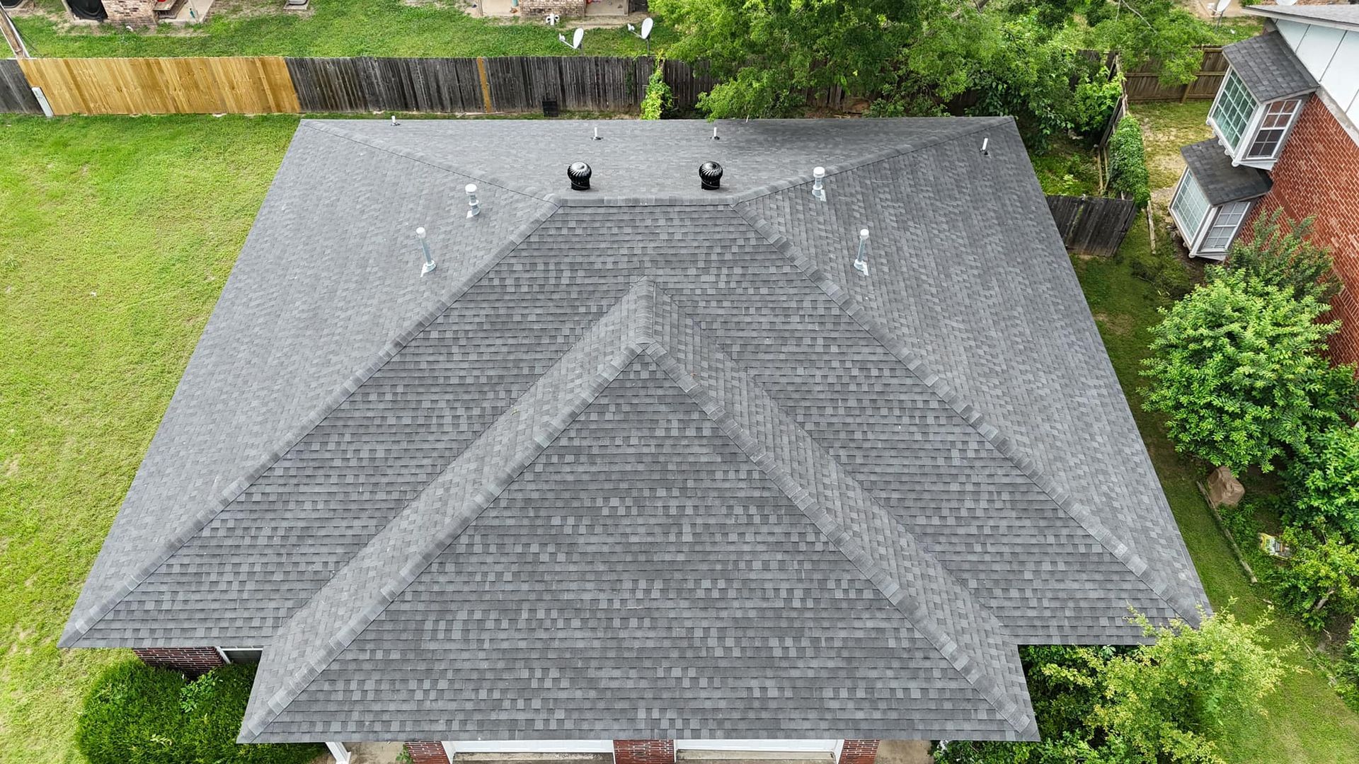 Aerial view of a gray shingled roof with a complex design; surrounded by green grass and trees.