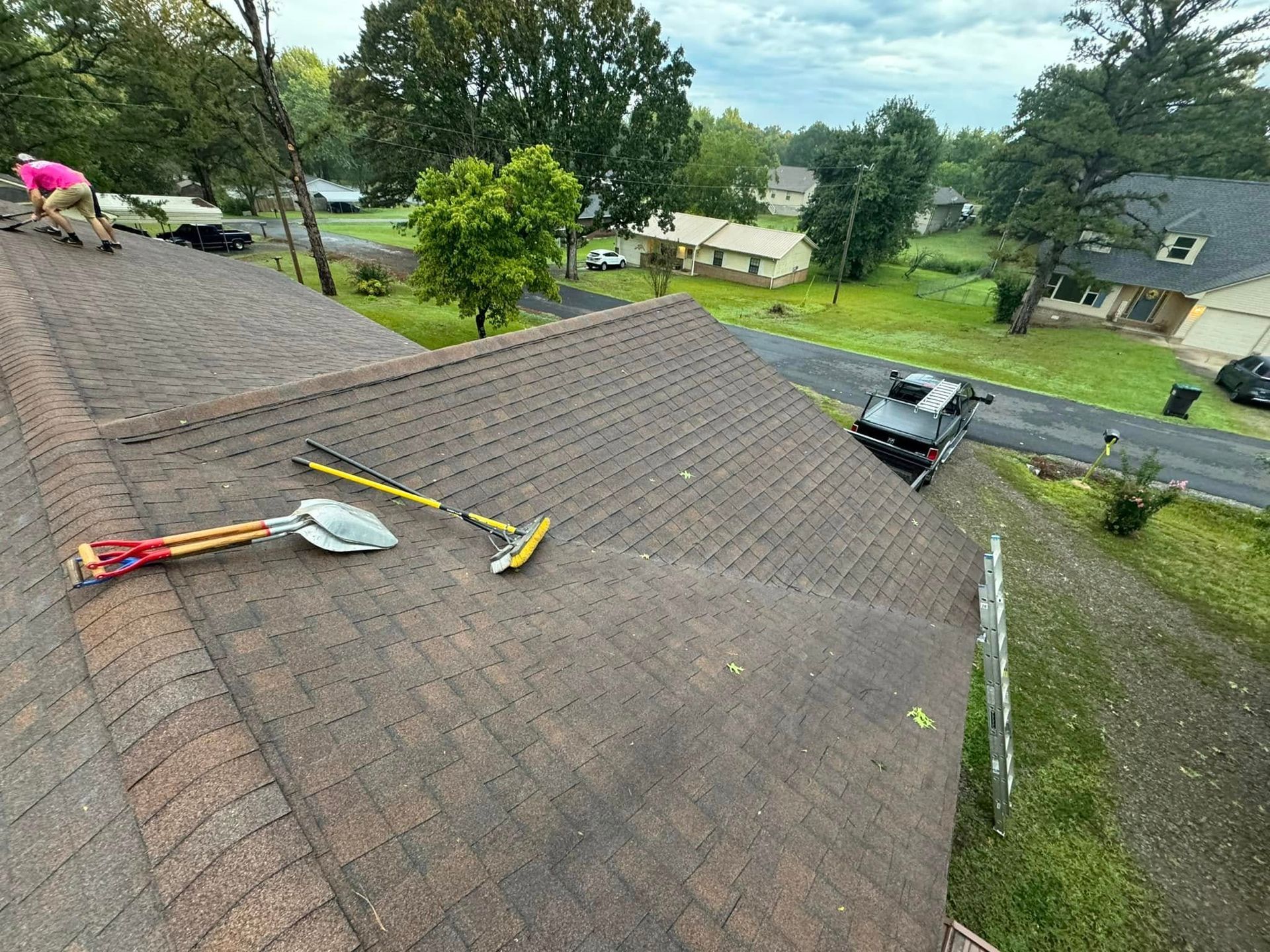 Roofing tools lie on a brown shingled roof, with workers in the background. Houses and street in view.