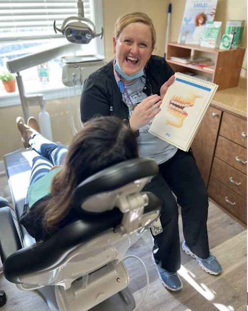 Dentist smiling and pointing at teeth diagram, explaining to a patient in a dental chair.