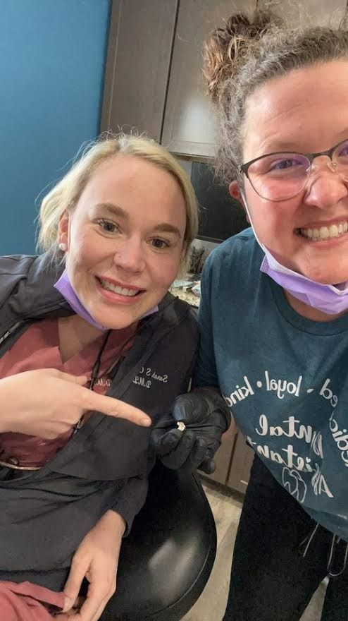Two women smiling, one points to a tooth. Dental office setting.