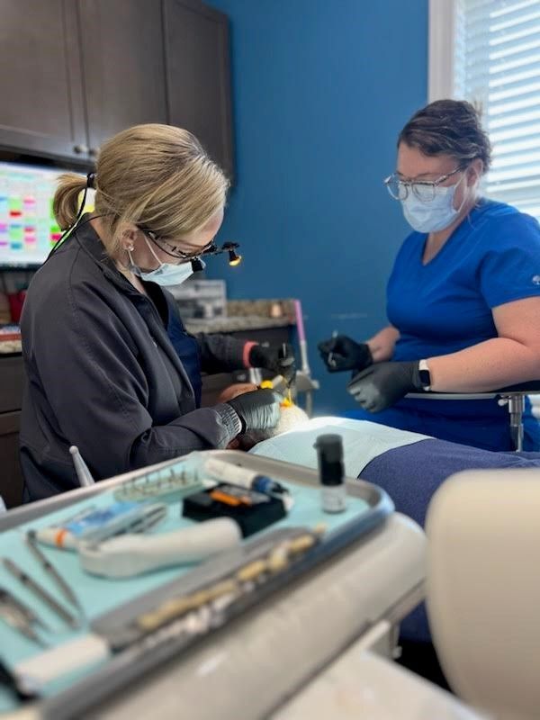 Two dental professionals perform a procedure in an office. One works on a patient, the other assists.