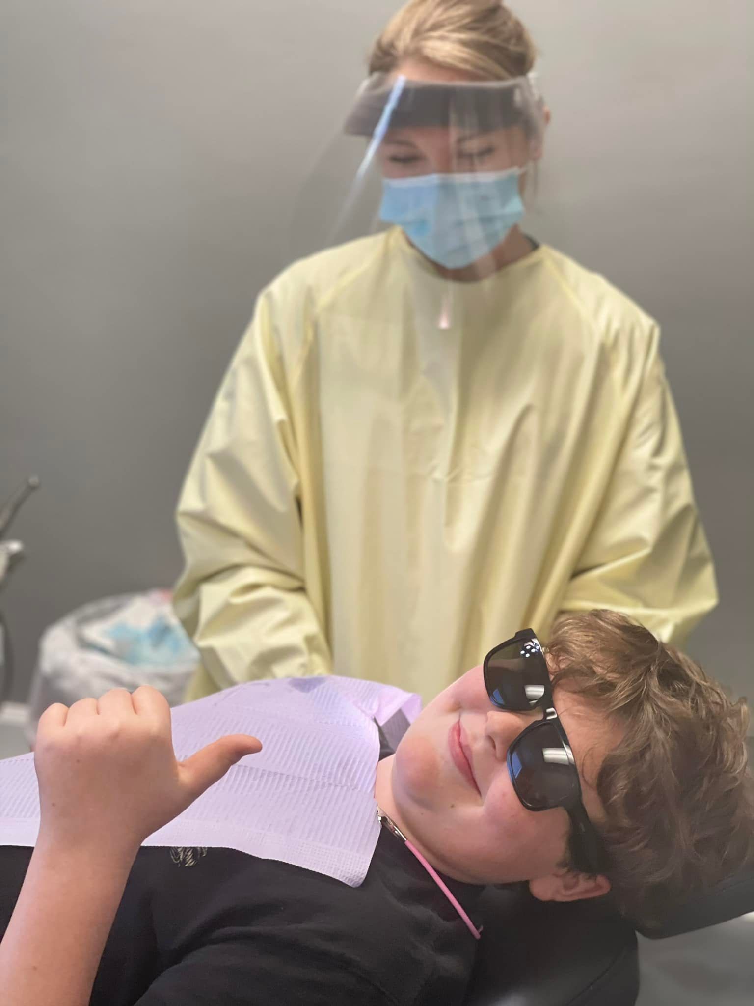Dentist in yellow gown, mask, and shield, tending to patient with sunglasses, in a dental office.