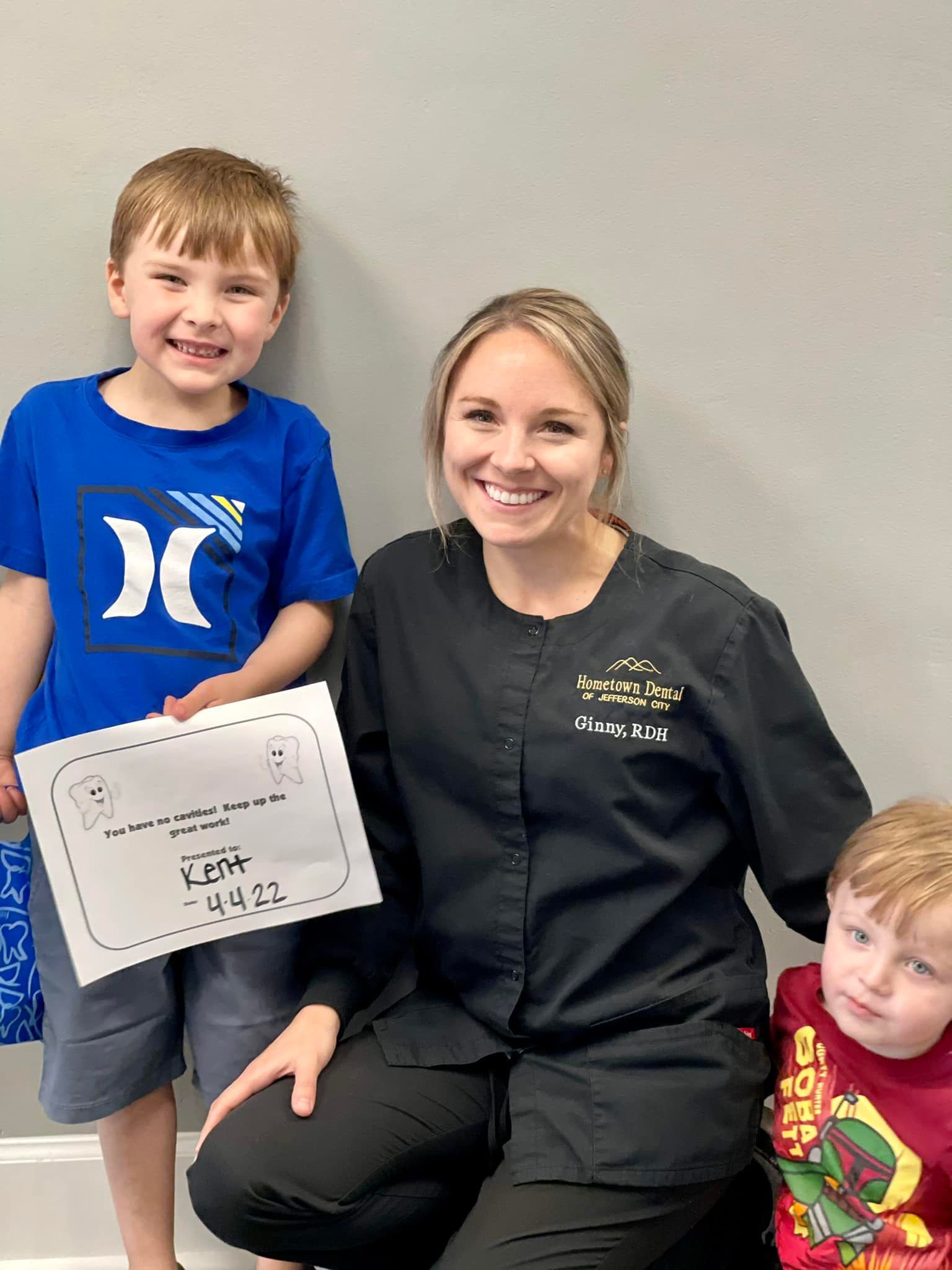 Woman in black scrubs with two smiling boys holding a certificate in a dentist's office.
