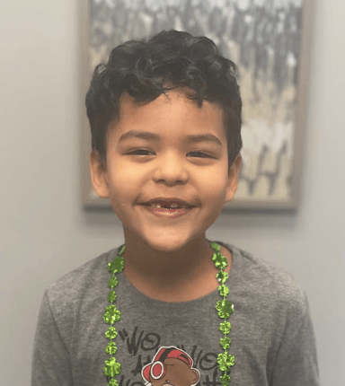 Smiling young boy with dark curly hair, missing a tooth, wearing a green shamrock necklace.