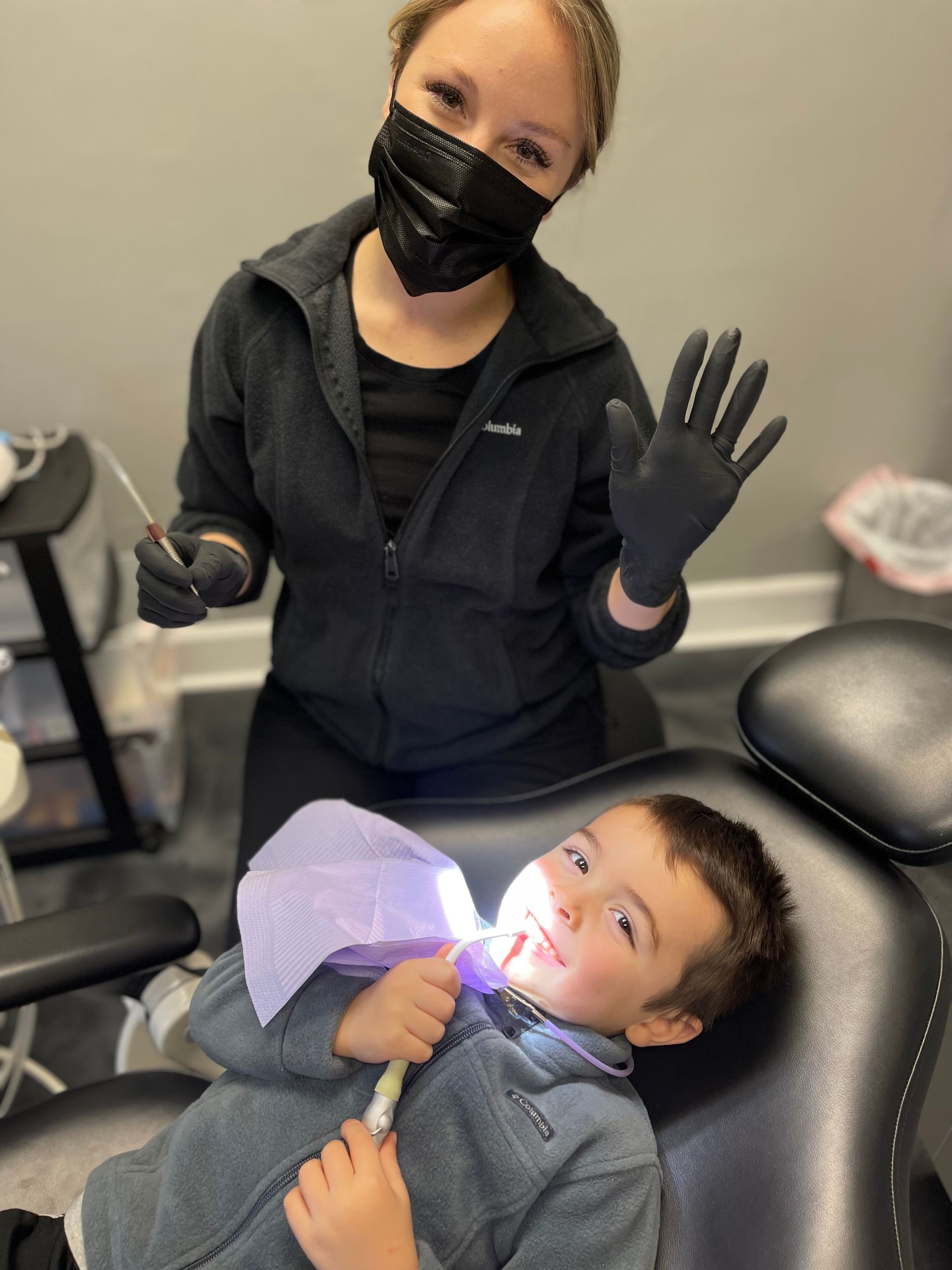 Dentist wearing mask and gloves waves at camera while treating a young boy in a dental chair.