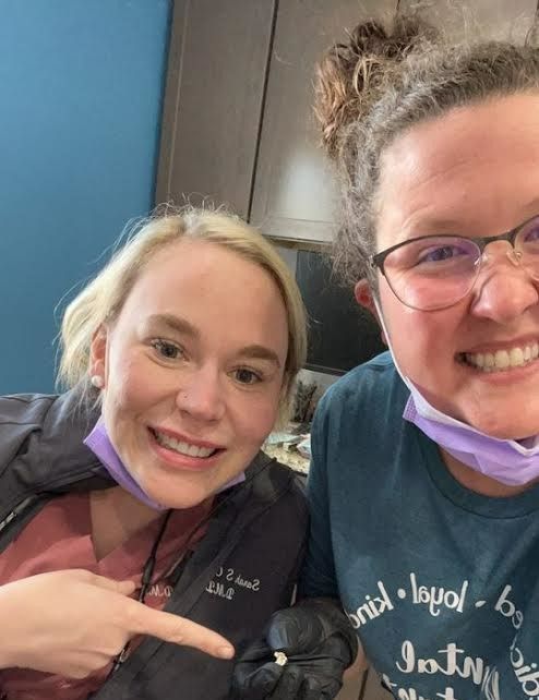 Two women smile, one pointing, in a dental office. One wears a teal shirt and glasses. The other, a black jacket.
