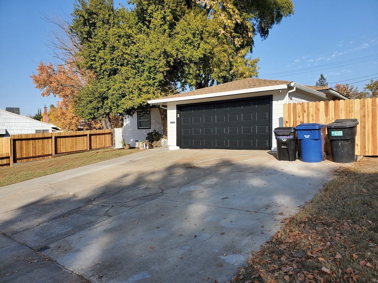 House with black garage door, driveway, wooden fence, and trash bins.