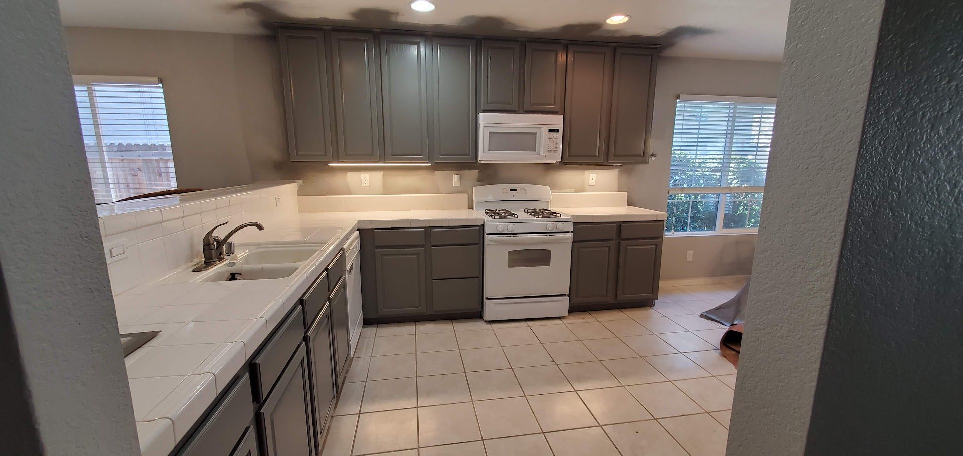 Kitchen with gray cabinets, white countertops, appliances, and tile floor; natural light from windows.