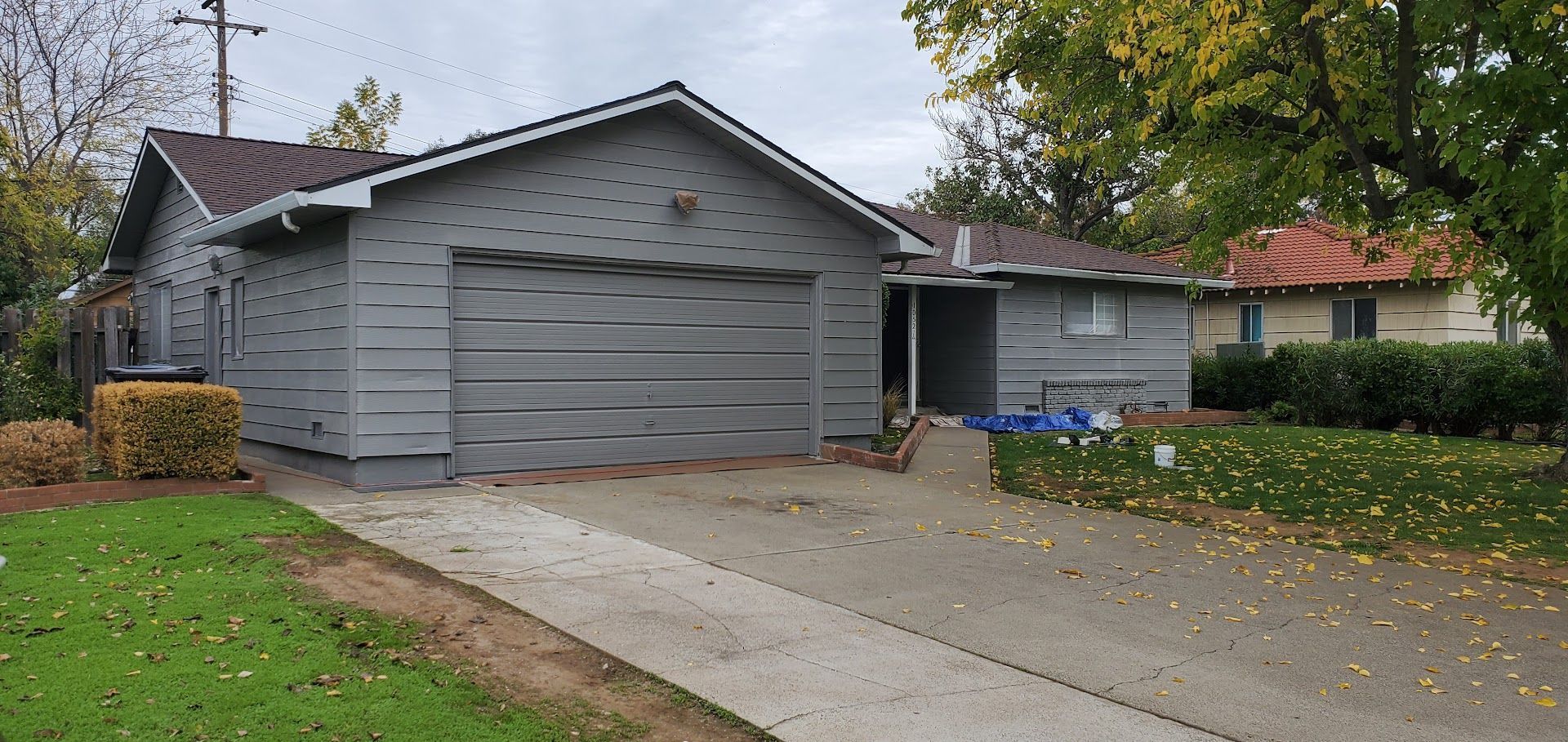 A gray house with a garage and driveway, grass on the left side, and a second house visible behind it.