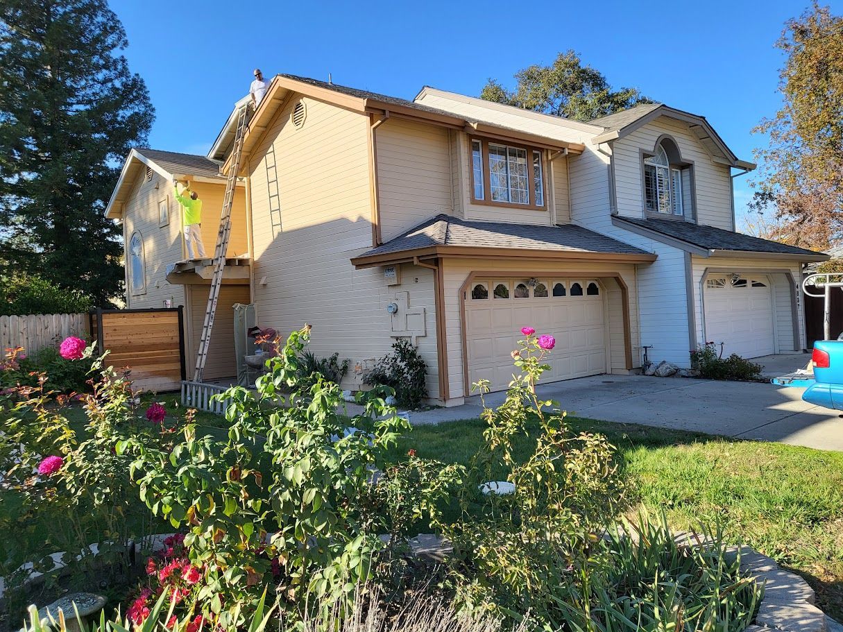 Two-story house with tan siding, brown trim, and a two-car garage. A ladder leans against the side.