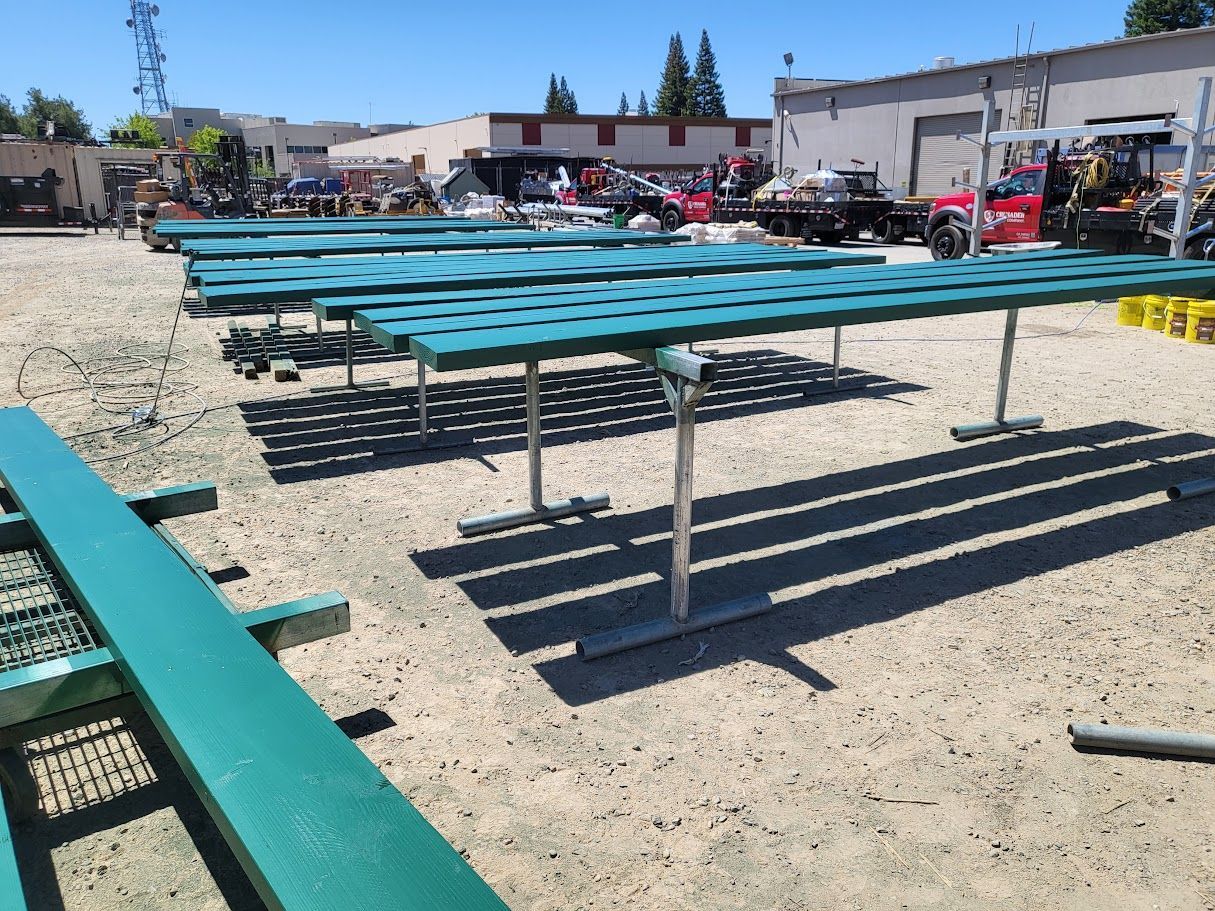 Green tables with holes on a gravel lot under a bright, sunny sky.