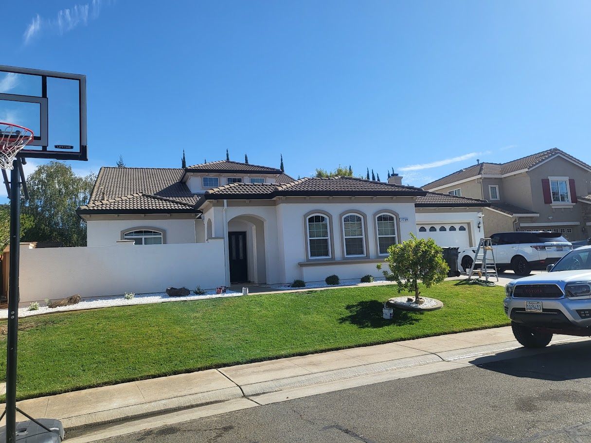 White house with tiled roof and green lawn, basketball hoop on the left, parked cars in front, blue sky.