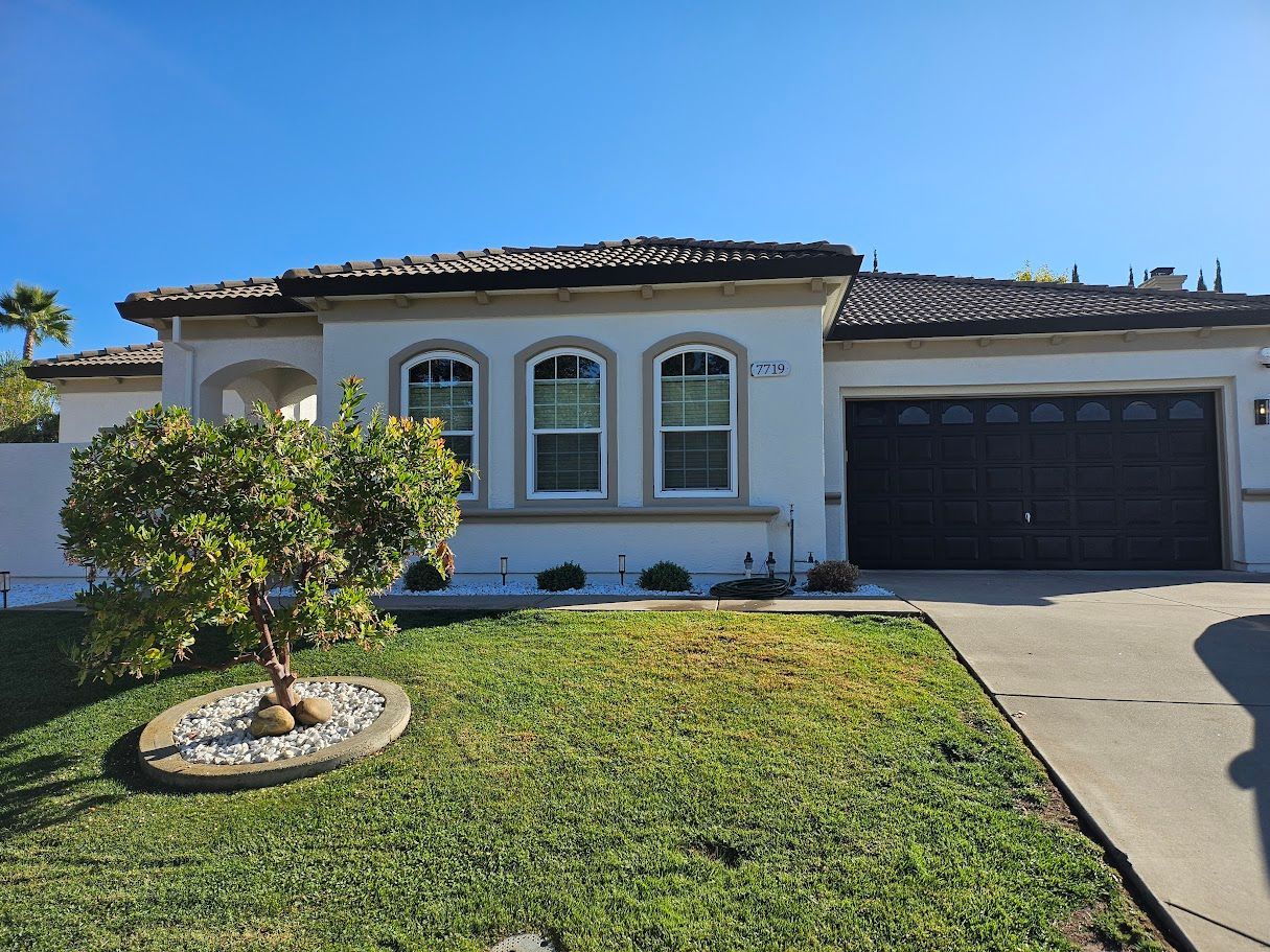 White house with a brown tile roof, three windows, and a dark garage door; green lawn and blue sky.