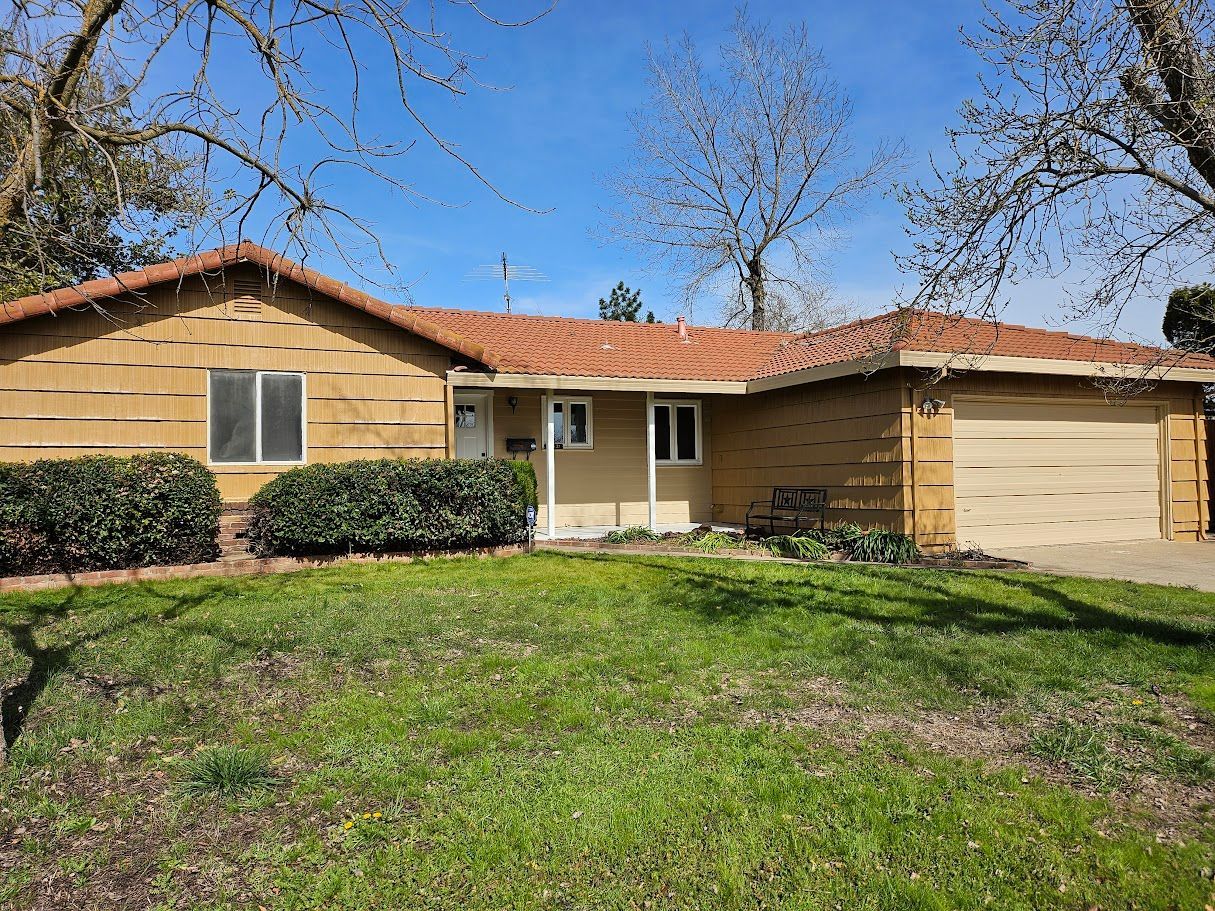 Single-story house with tan siding, red tile roof, and attached garage; green lawn in front.
