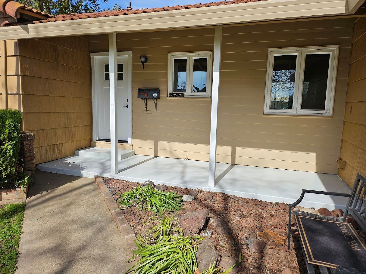 Tan house with covered porch, white door and window frames. Concrete porch and pathway with bench and garden in front.