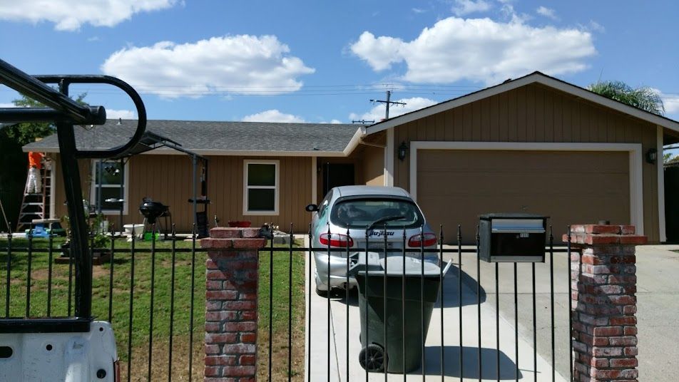 Tan house with a gray roof, a parked silver car, and a black fence on a sunny day.
