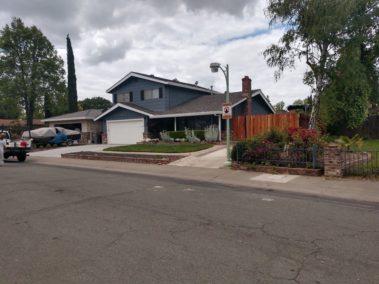 Two-story blue house with white garage door, brown fence, and landscaping. Overcast sky.