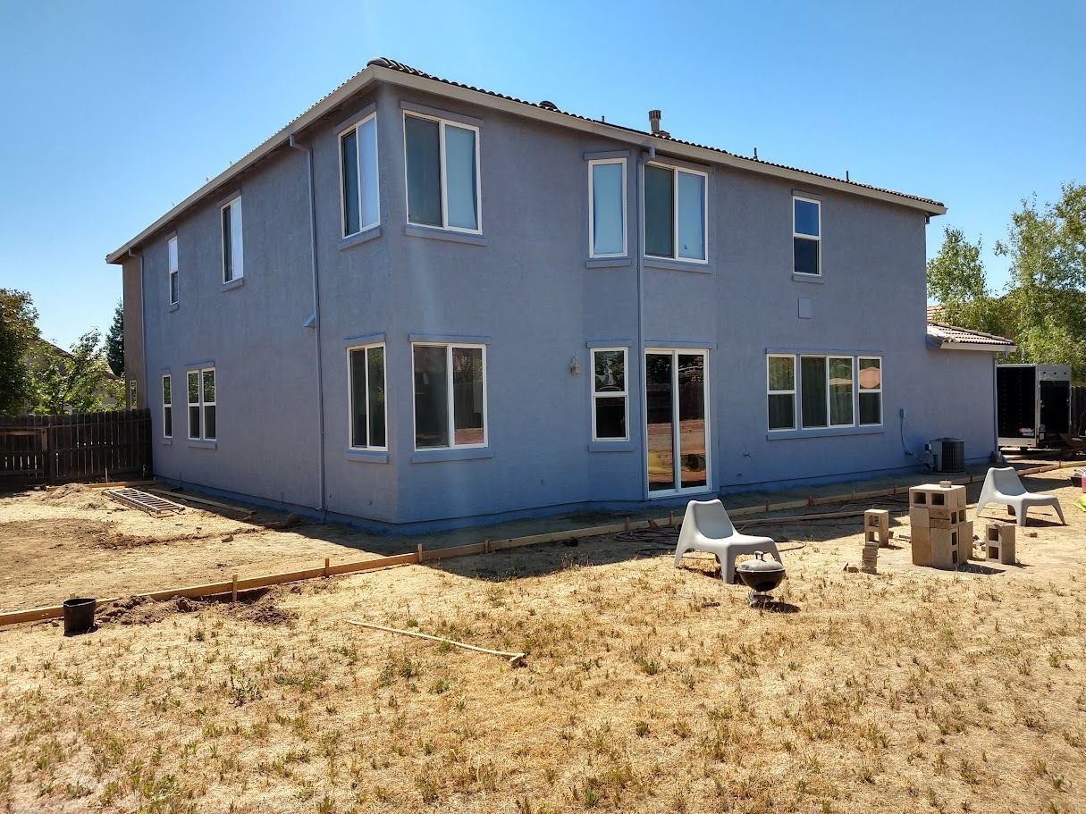 Back of a two-story blue house with multiple windows and a yard with dry grass and chairs.