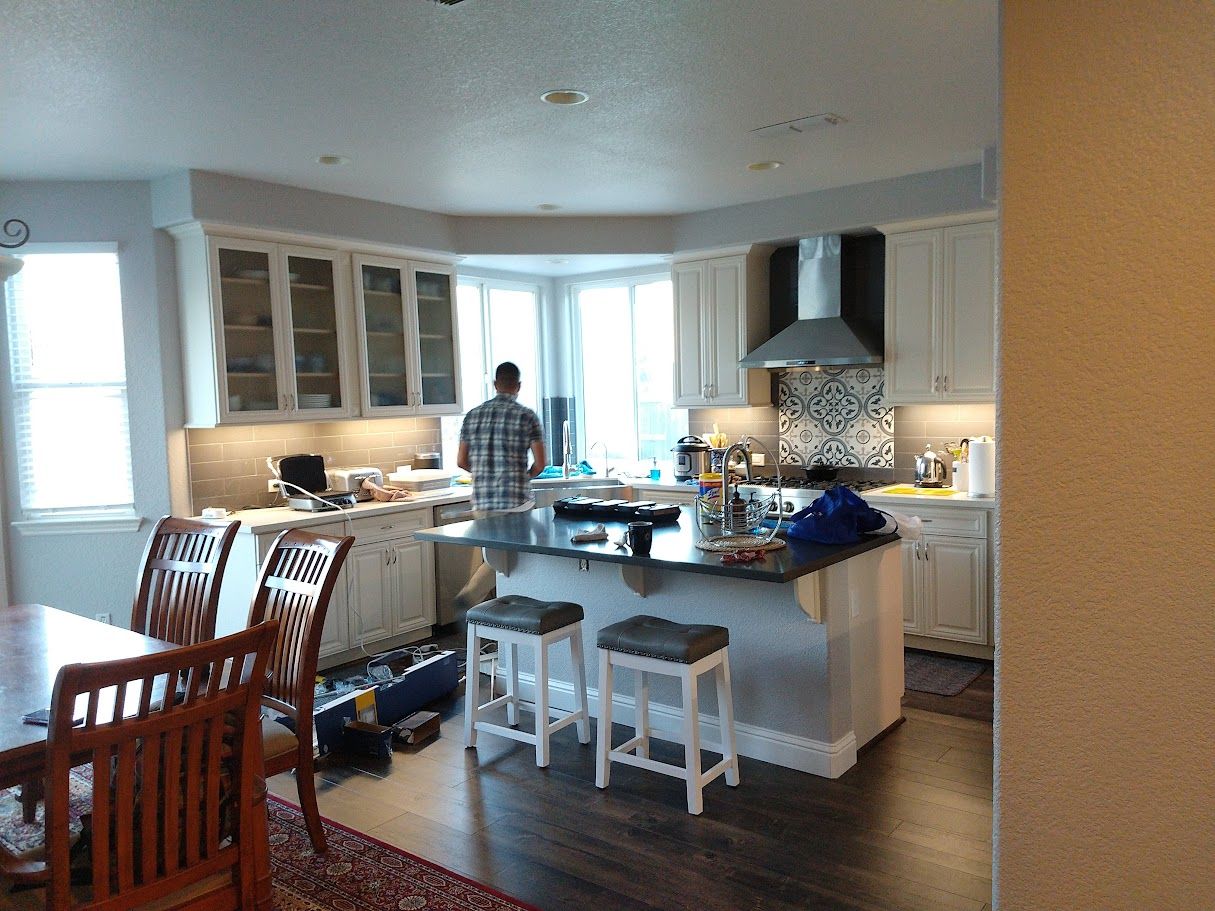 Kitchen with white cabinets, island with stools, a person standing by a window, and dining table.