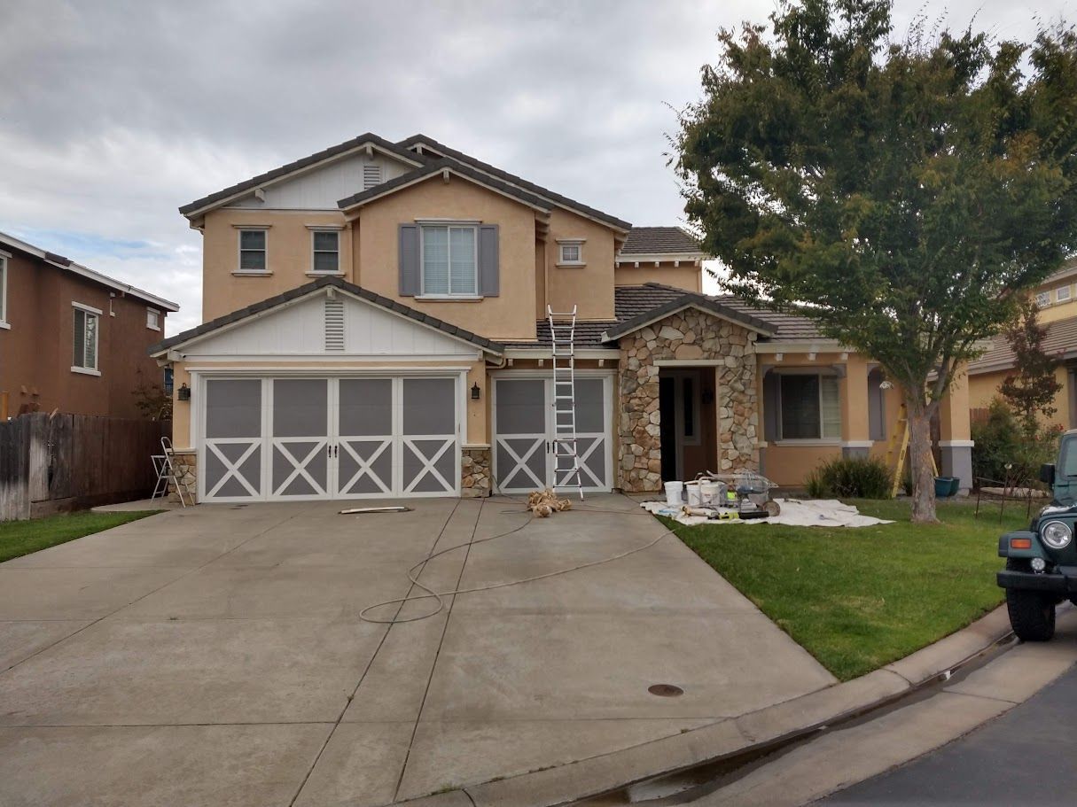 Two-story house with tan stucco, stone accents, white garage doors, and a concrete driveway.
