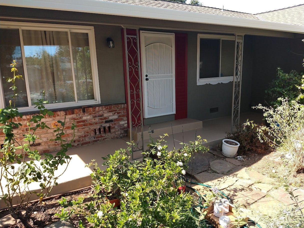 A house exterior with exposed brick, gray siding, and a white security door. Plants in the foreground.