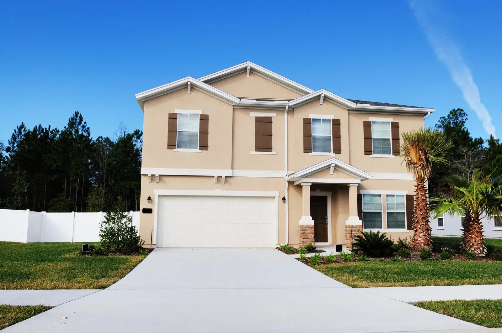 Two-story beige house with white garage door and brown shutters against a blue sky.