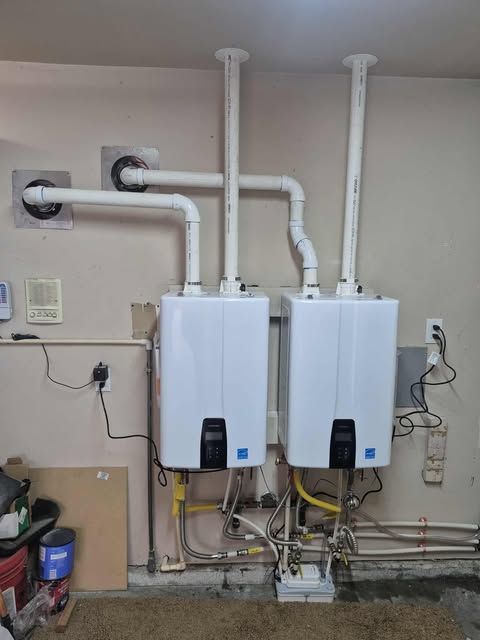 Two white tankless water heaters mounted on a beige wall with white PVC venting.