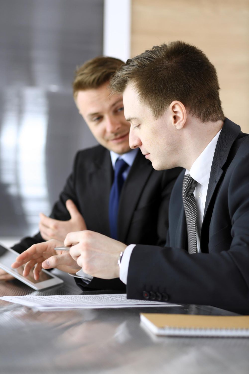 Two professionals in suits reviewing documents and a tablet together at an office desk.