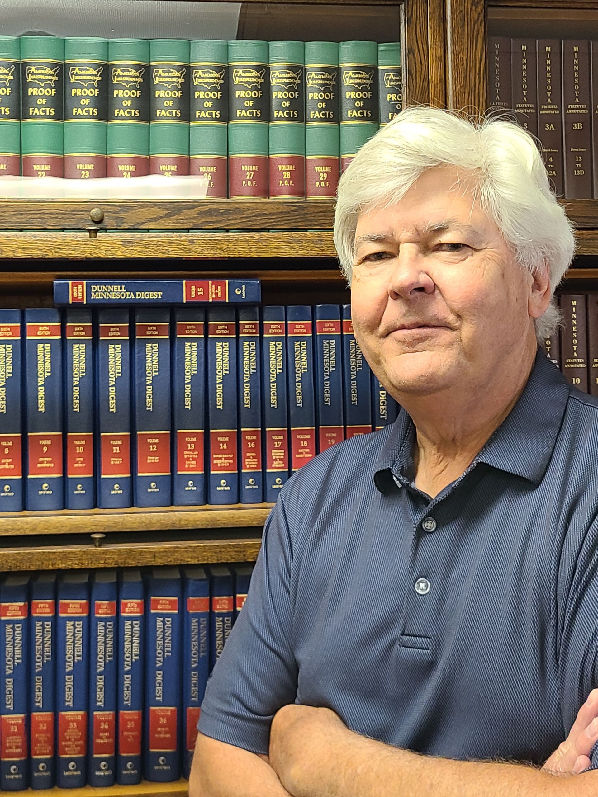 A person with light hair stands with arms crossed in front of a wall of bookshelves filled with law books.