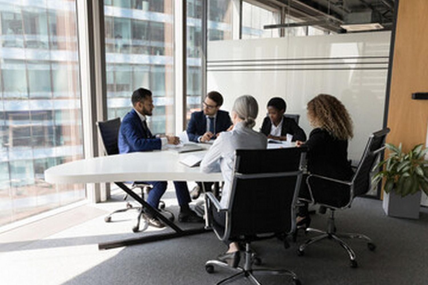 A diverse professional team sits around a sleek white table in a sunlit office, engaged in a collaborative meeting.