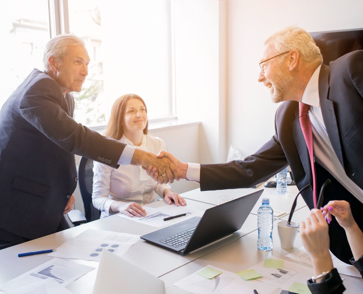 Two professionals in suits shake hands across a conference table while a colleague looks on in a bright, modern office.